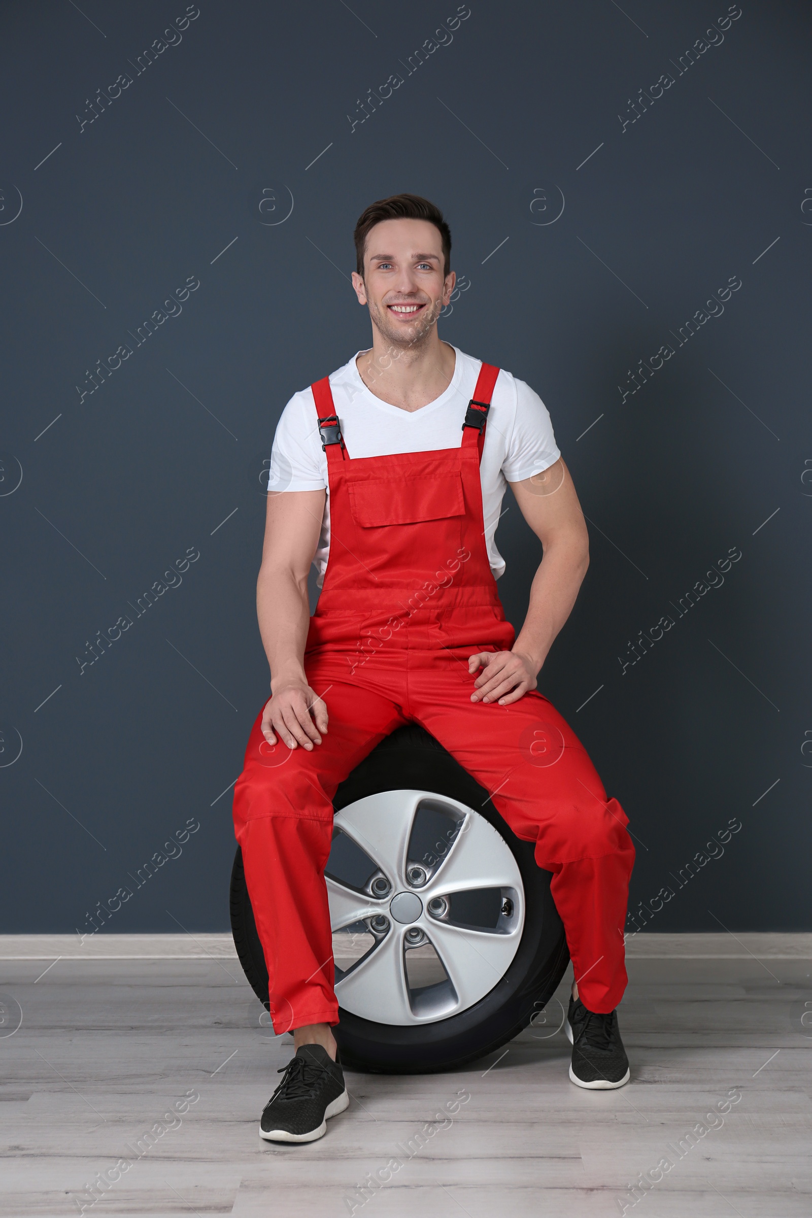 Photo of Young mechanic in uniform sitting on car tire near dark wall