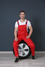 Young mechanic in uniform sitting on car tire near dark wall