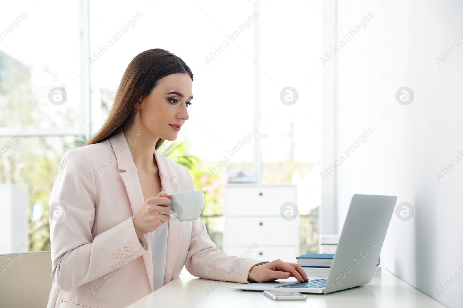 Image of Young woman drinking coffee while working on laptop in office