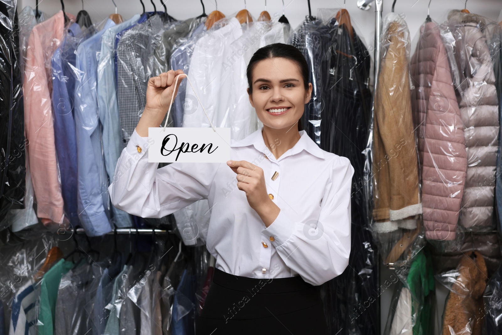 Photo of Dry-cleaning service. Happy worker holding Open sign near rack with clothes indoors