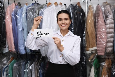 Photo of Dry-cleaning service. Happy worker holding Open sign near rack with clothes indoors