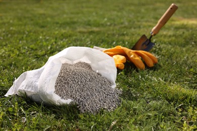 Photo of Granulated fertilizer in sack, shovel and gloves on green grass outdoors, closeup