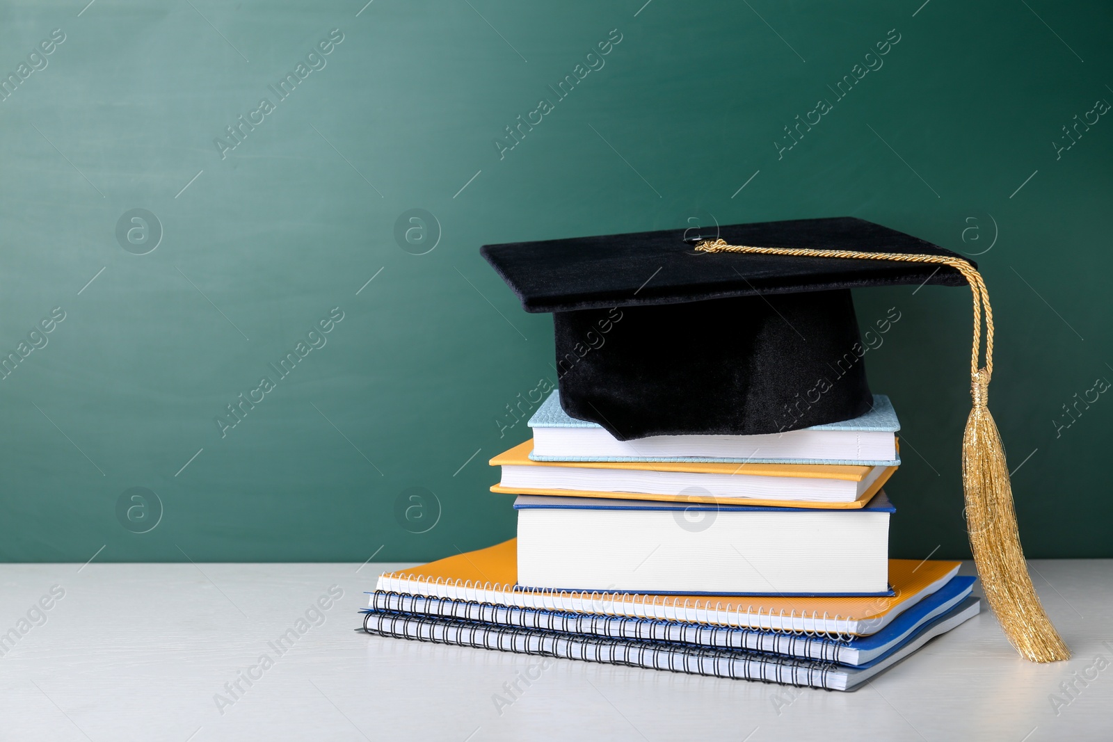 Photo of Graduation hat with books and notebooks on table near chalkboard. Space for text