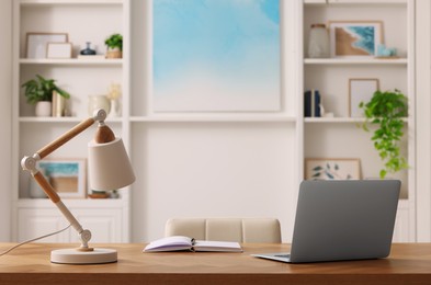 Photo of Interior design. Cosy workplace with laptop and lamp on wooden table near abstract picture between shelves