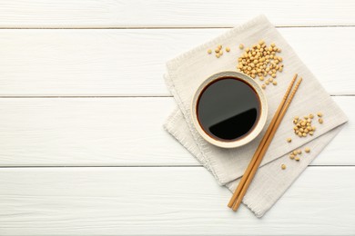 Tasty soy sauce in bowl, chopsticks and soybeans on white wooden table, top view. Space for text