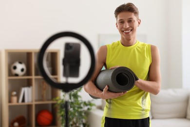 Photo of Smiling sports blogger holding yoga mat while streaming online fitness lesson at home