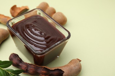 Photo of Bowl of tasty tamarind sauce, leaves and fresh pods on light green table. Space for text