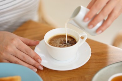 Photo of Breakfast. Woman pouring cream into cup of coffee at wooden table, closeup