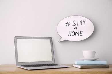 Photo of Laptop, cup and speech bubble with hashtag STAY AT HOME indoors. Message to promote self-isolation during COVID‑19 pandemic