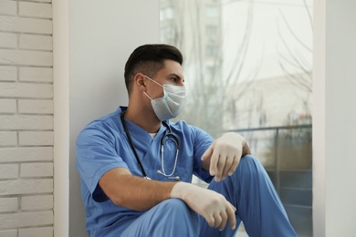 Photo of Sad doctor near window indoors. Stress of health care workers during coronavirus pandemic