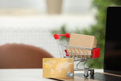 Photo of Online payment concept. Small shopping cart with bank card, box and laptop on white table