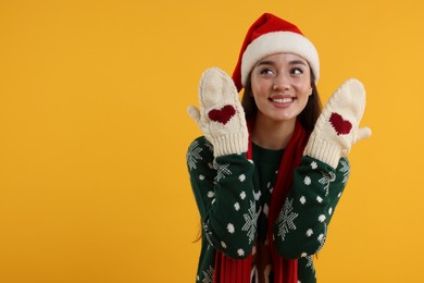 Photo of Happy young woman in Christmas sweater, Santa hat and knitted mittens on orange background. Space for text