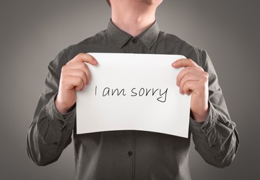 Image of Apology. Man holding card with phrase I Am Sorry on grey background, closeup