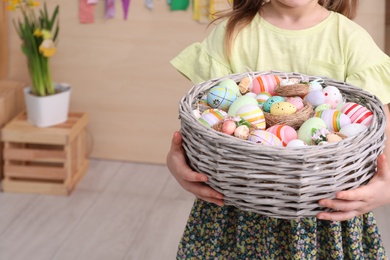 Photo of Little girl holding wicker basket full of Easter eggs indoors, closeup. Space for text