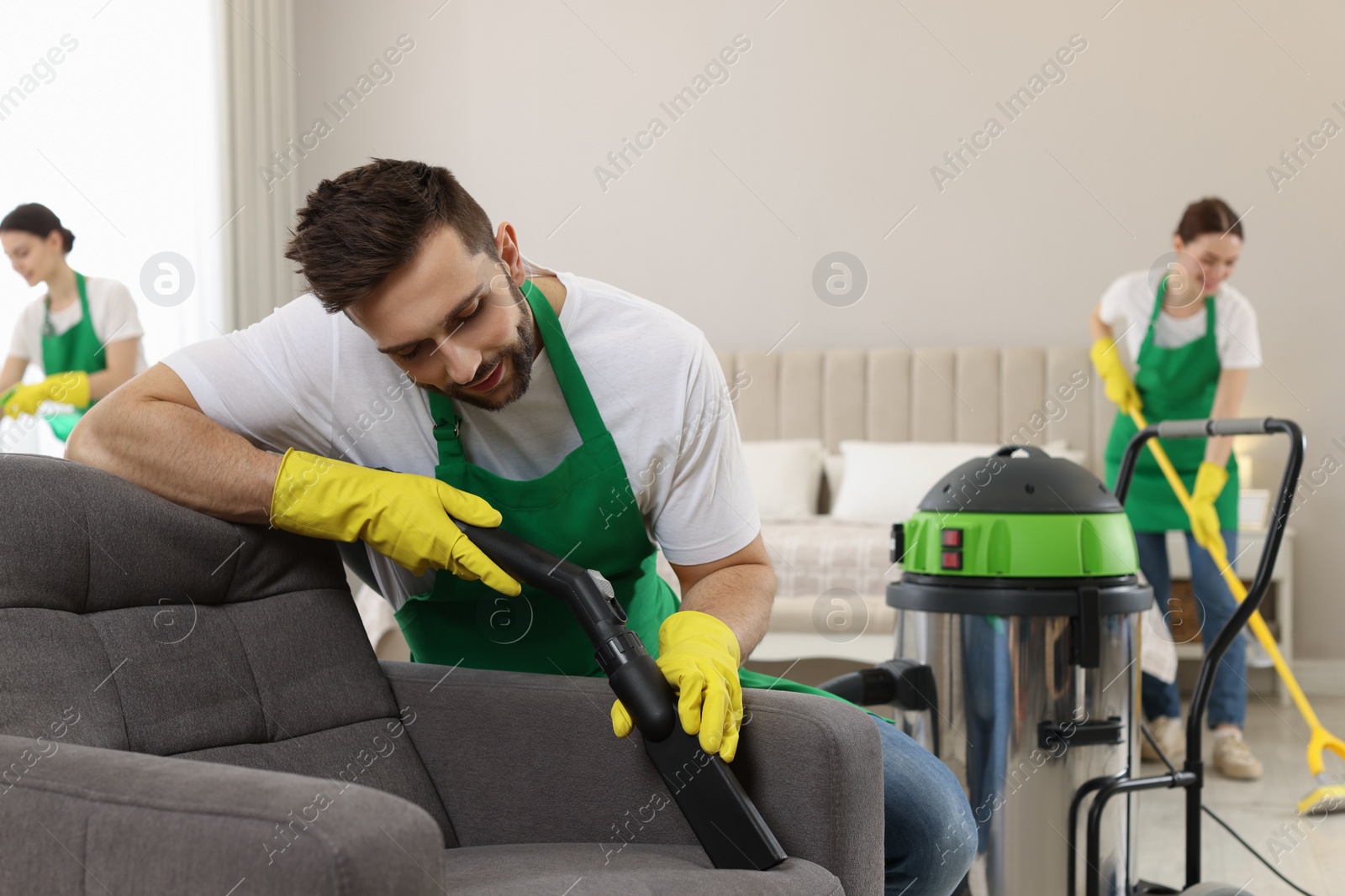 Photo of Professional janitor in uniform vacuuming armchair indoors