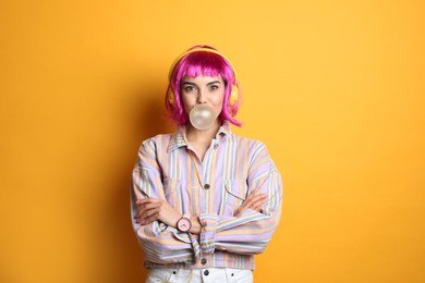 Fashionable young woman in colorful wig with headphones blowing bubblegum on yellow background