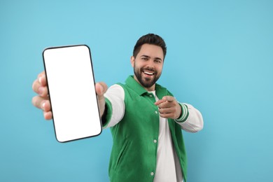 Photo of Young man showing smartphone in hand and pointing at camera on light blue background