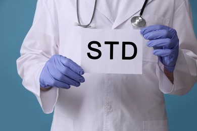 Photo of Doctor holding sheet of paper with abbreviation STD on light blue background, closeup