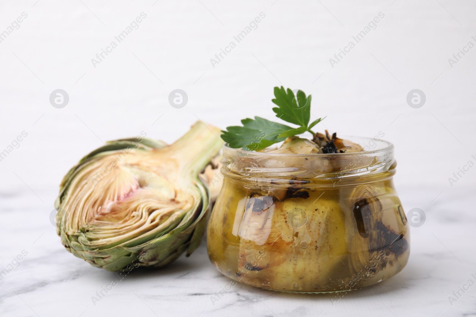 Photo of Jar of delicious artichokes pickled in olive oil and fresh vegetables on white marble table