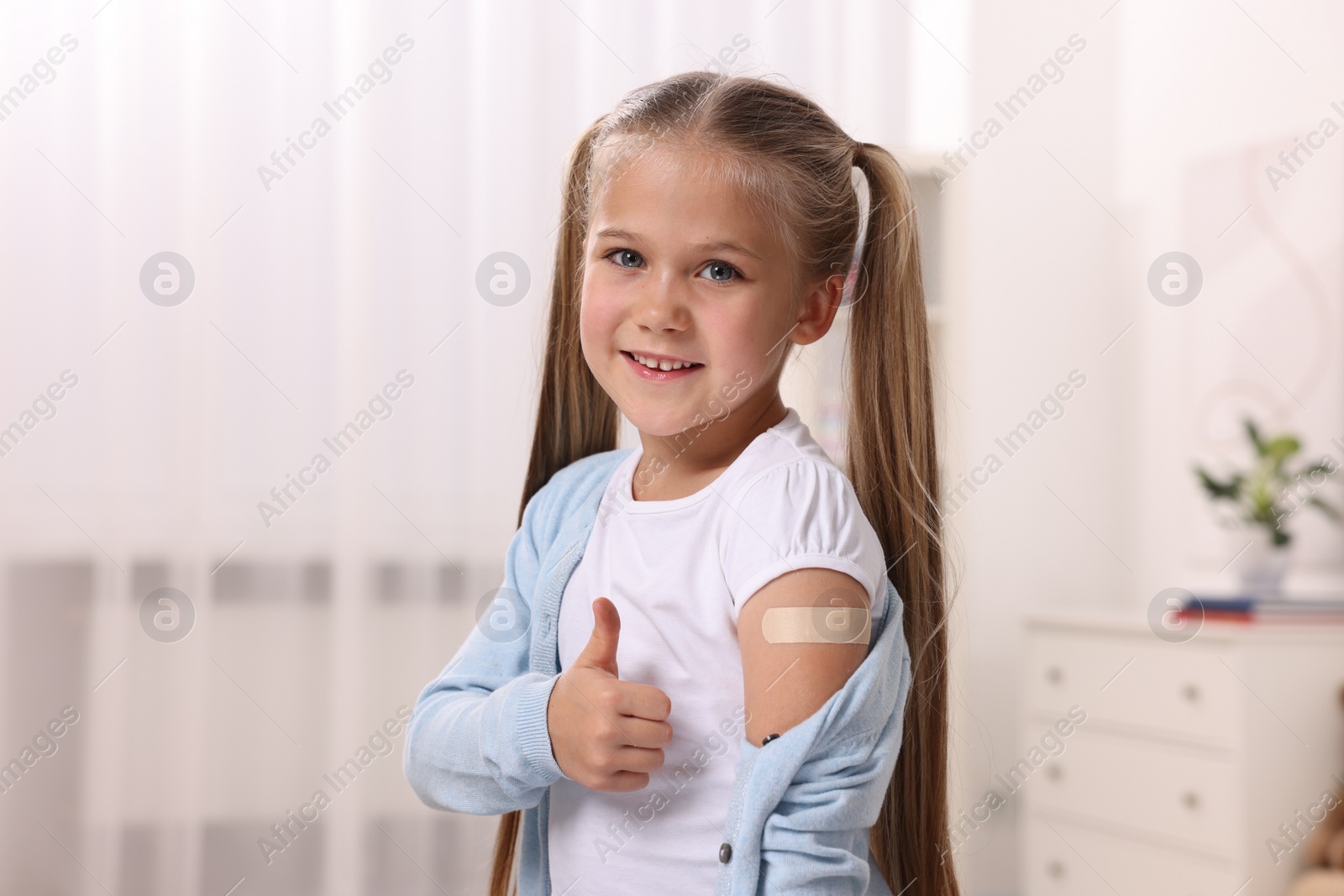 Photo of Happy girl with sticking plaster on arm after vaccination showing thumbs up indoors