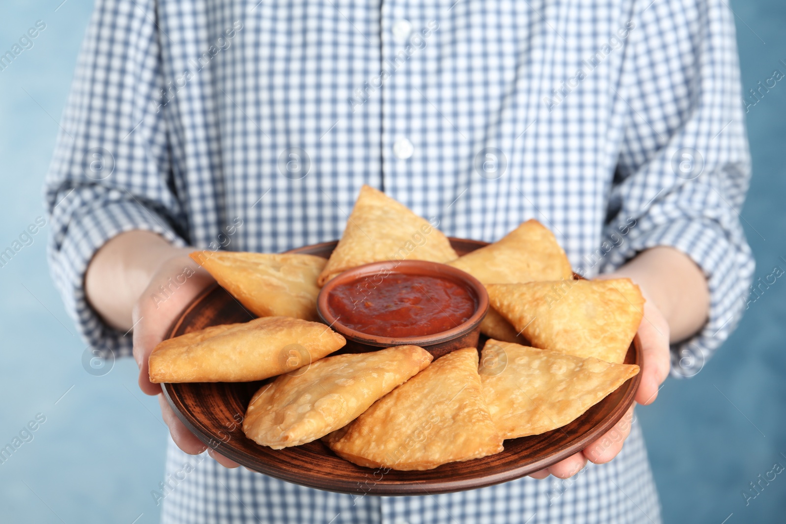 Photo of Woman holding plate with delicious samosas and sauce on light blue background, closeup