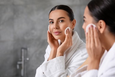 Photo of Beautiful woman removing makeup with cotton pads near mirror in bathroom