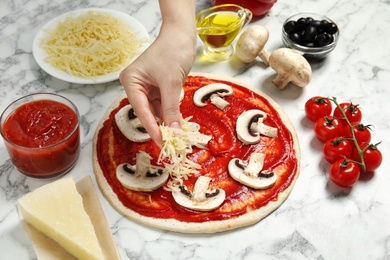 Woman adding grated cheese to unbaked pizza on marble table, closeup