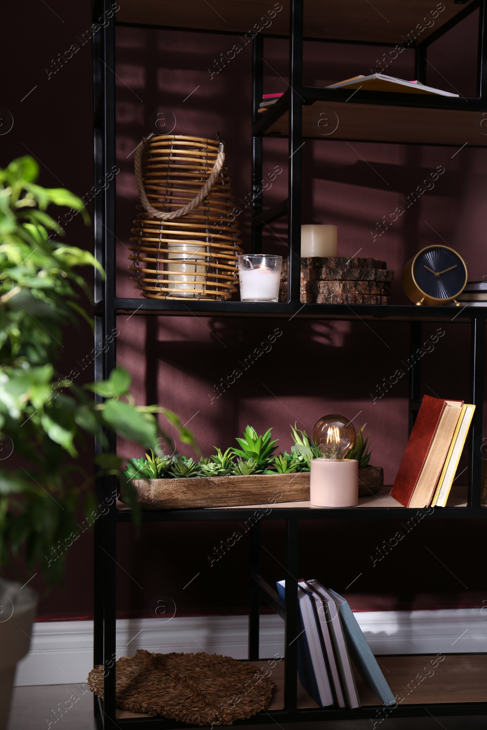 Photo of Stylish wicker candle holder, night lamp with incandescent bulb and decor on shelving unit near brown wall