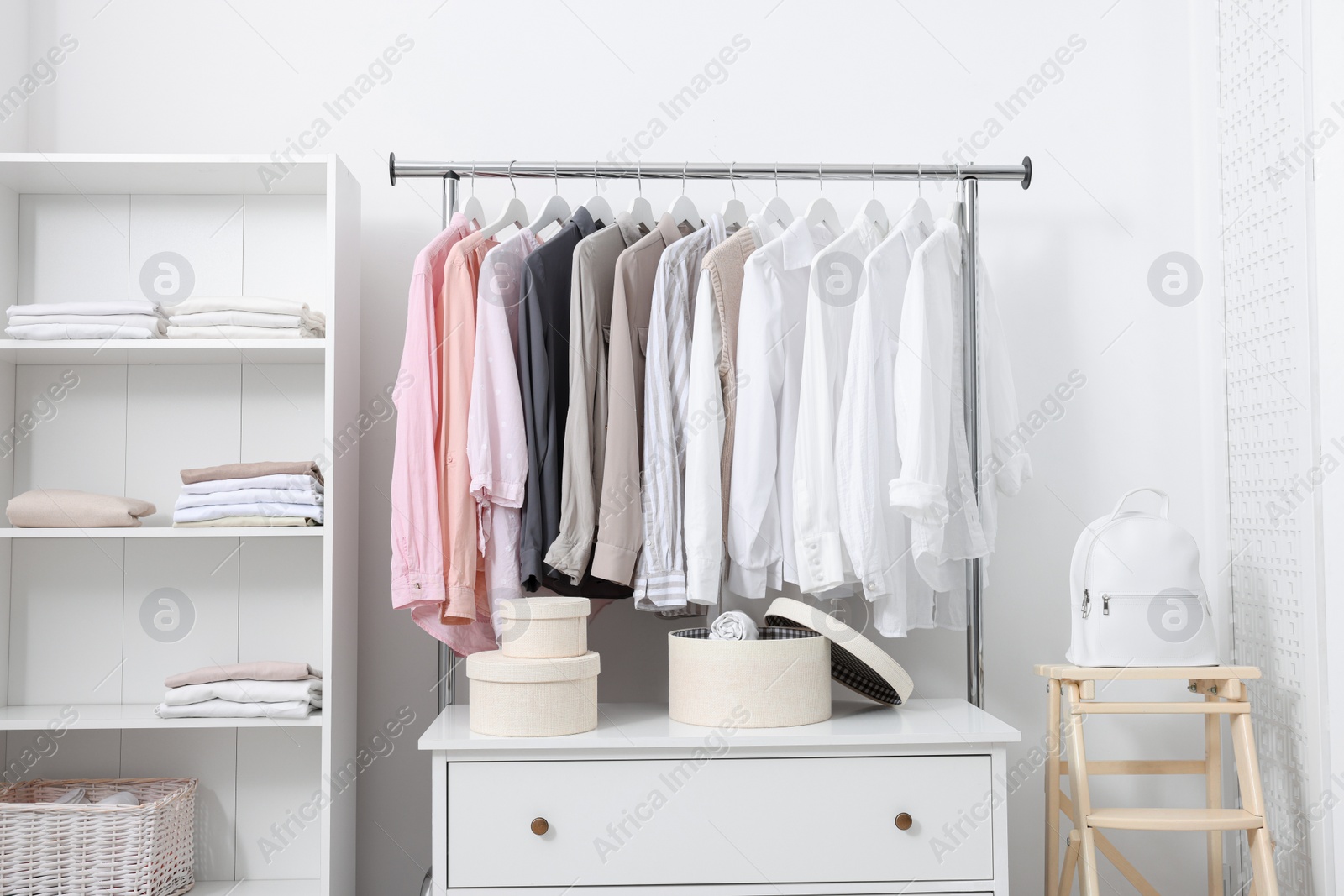 Photo of Wardrobe organization. Rack with different stylish clothes, chest of drawers and shelving unit near white wall indoors