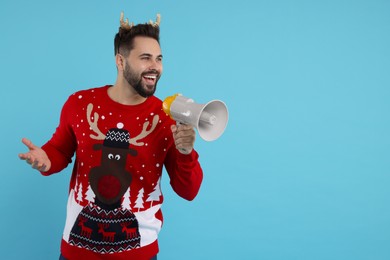 Photo of Happy young man in Christmas sweater and reindeer headband shouting in megaphone on light blue background. Space for text