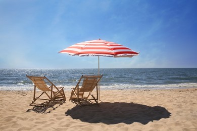 Photo of Deck chairs near red and white striped beach umbrella on sandy seashore
