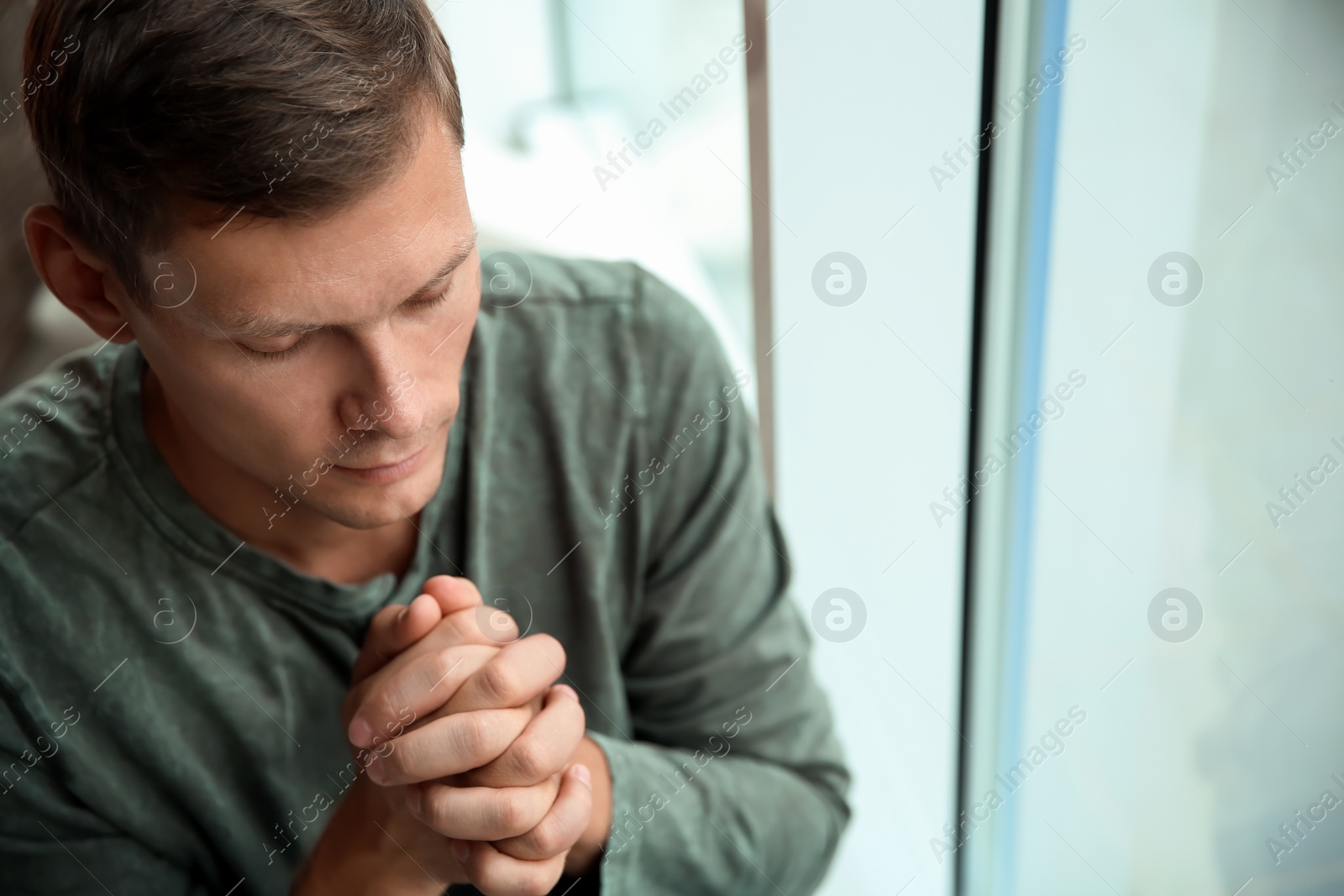 Photo of Man with hands clasped together for prayer near window. Space for text