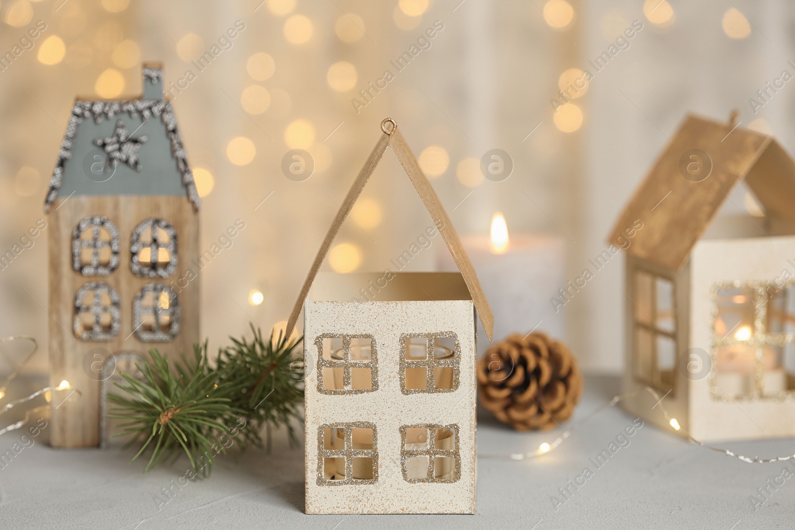 Photo of House shaped holders with burning candles on grey table against blurred Christmas lights