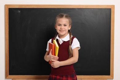 Photo of Happy schoolgirl with colorful books near blackboard