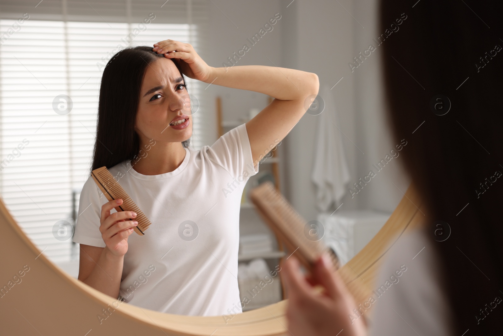 Photo of Emotional woman with comb examining her hair and scalp near mirror in bathroom. Dandruff problem