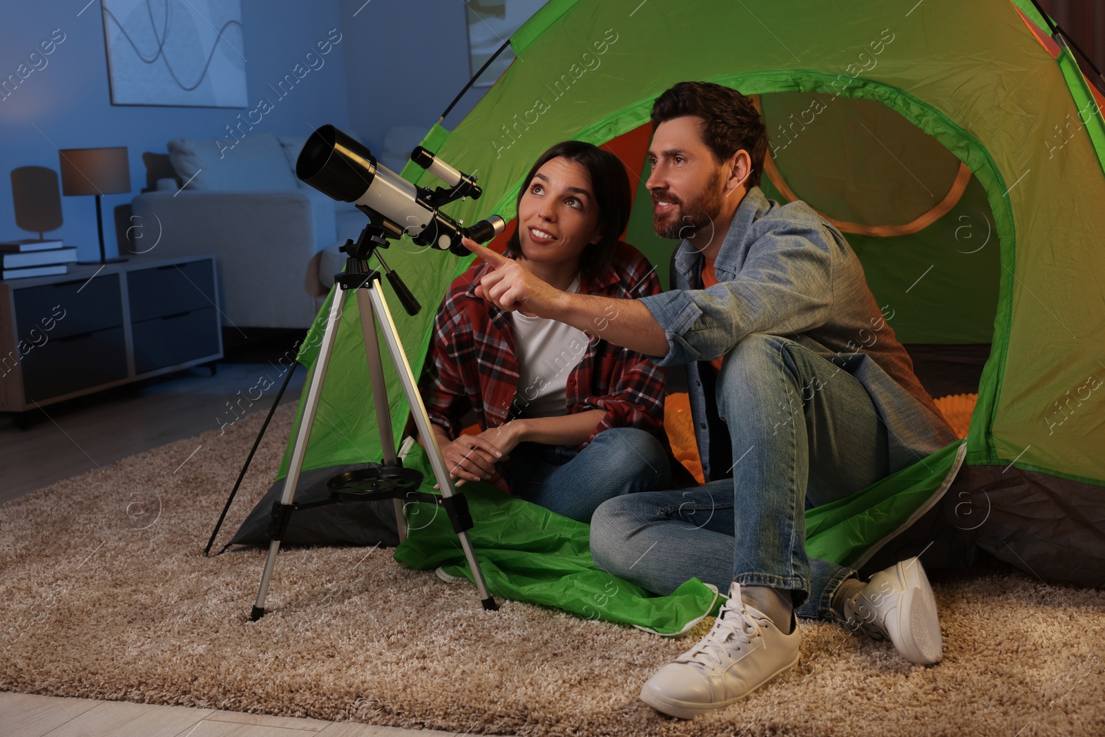 Photo of Happy couple using telescope to look at stars while sitting in camping tent indoors