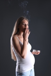 Photo of Young pregnant woman smoking cigarette on dark background. Harm to unborn baby