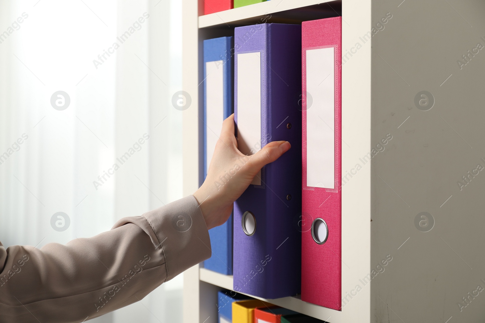 Photo of Woman taking binder office folder from shelving unit indoors, closeup