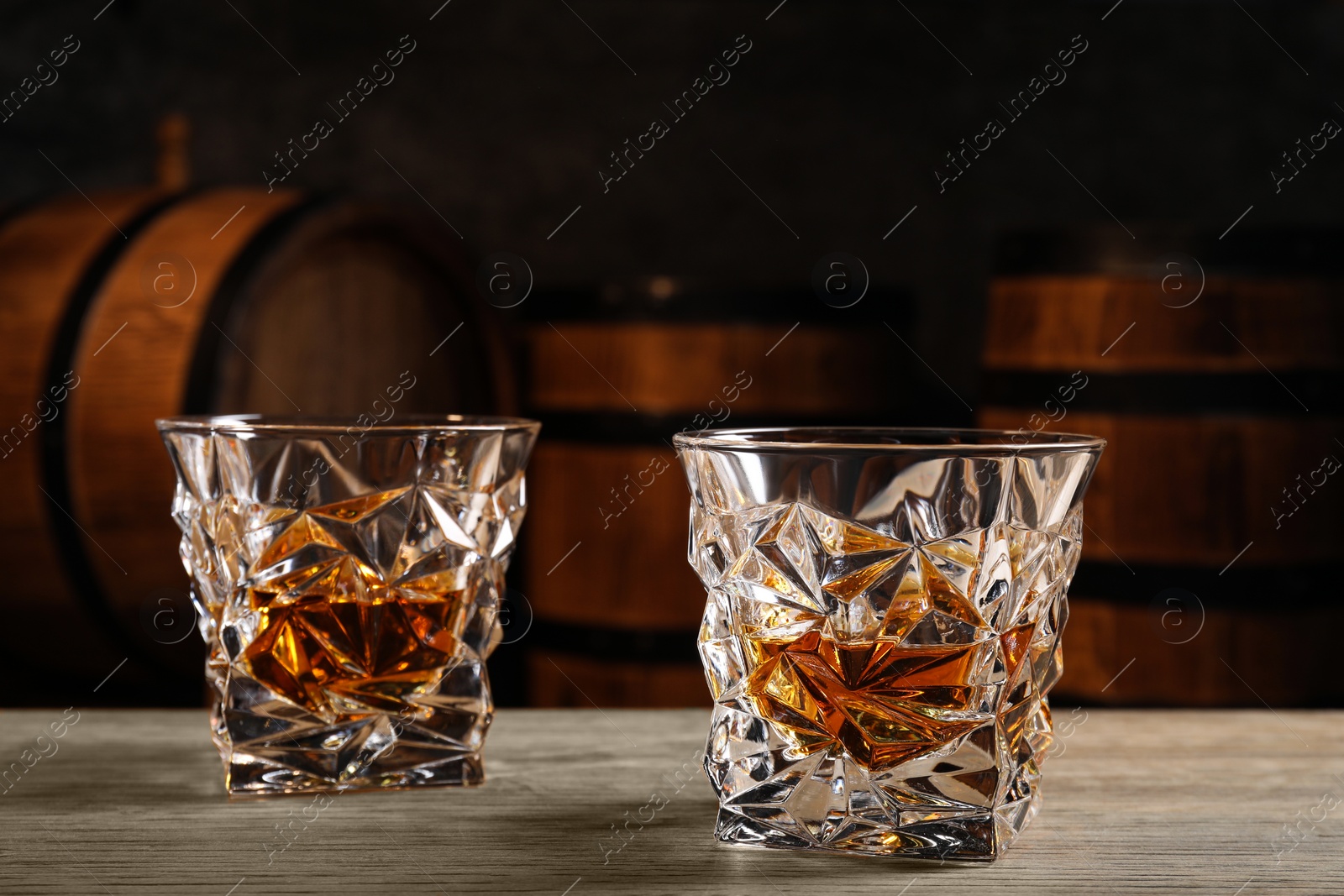 Photo of Glasses with whiskey on table against wooden barrels, closeup