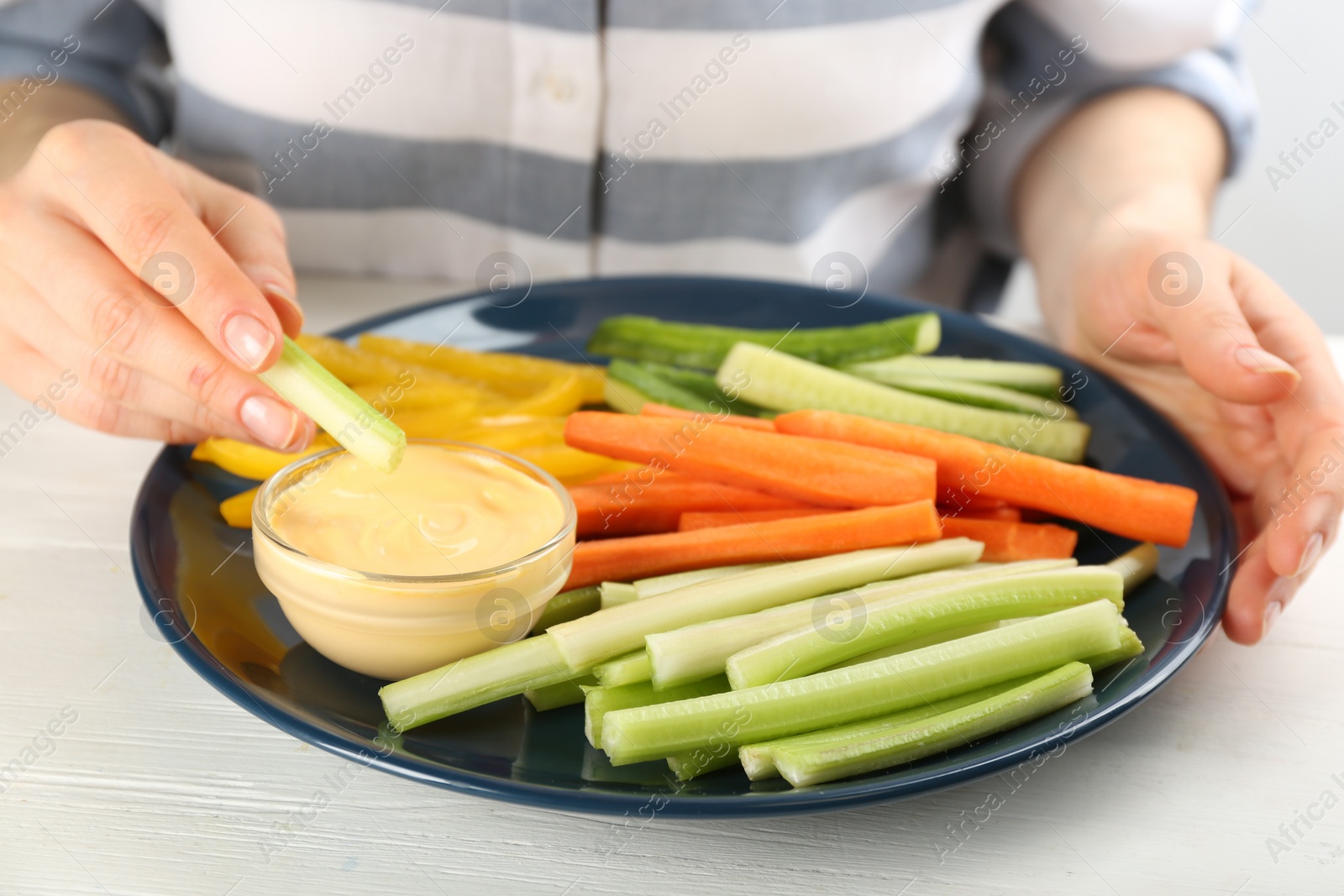 Photo of Woman dipping celery stick in sauce at white wooden table, closeup