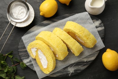 Photo of Delicious cake roll, lemons, mint and powdered sugar on black table, flat lay