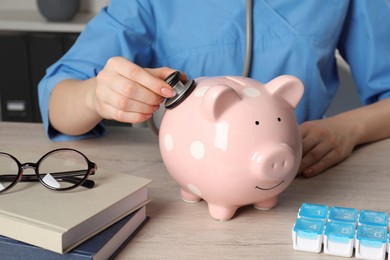 Doctor with stethoscope and piggy bank at wooden table in hospital, closeup. Medical insurance