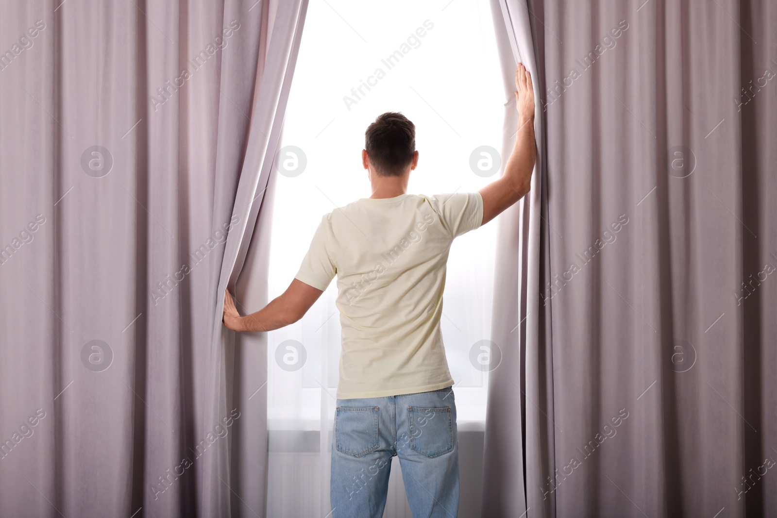 Photo of Man opening window curtains at home, back view