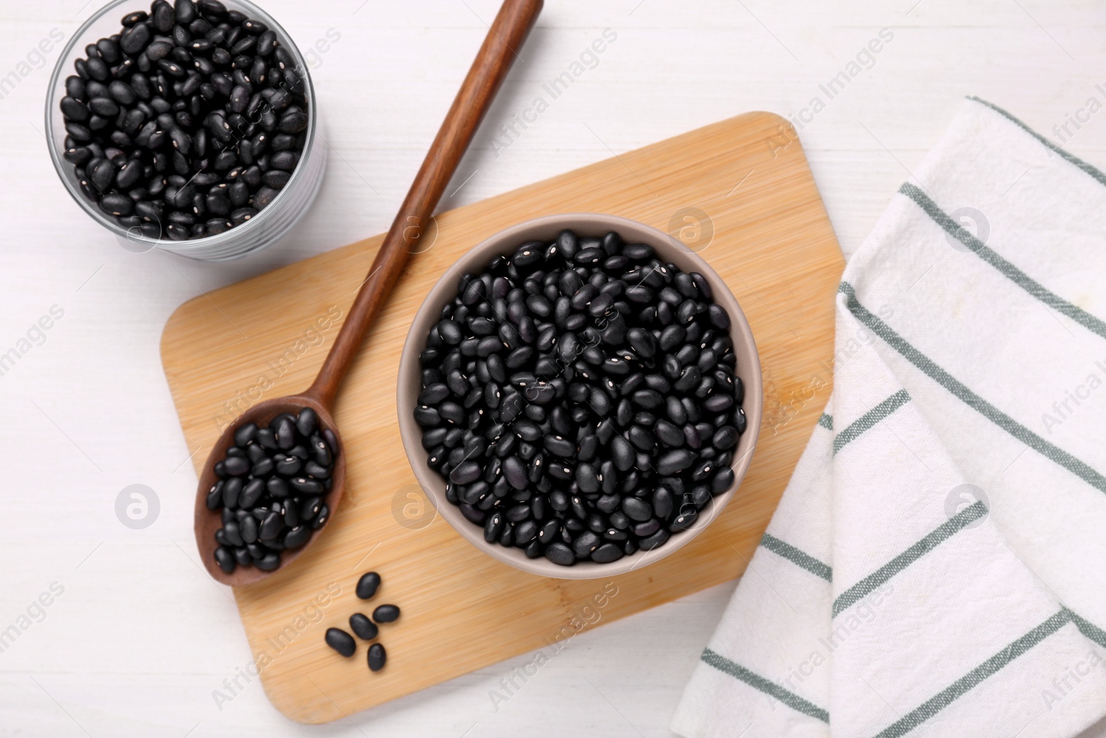 Photo of Bowl and spoon of raw black beans on white wooden table, flat lay