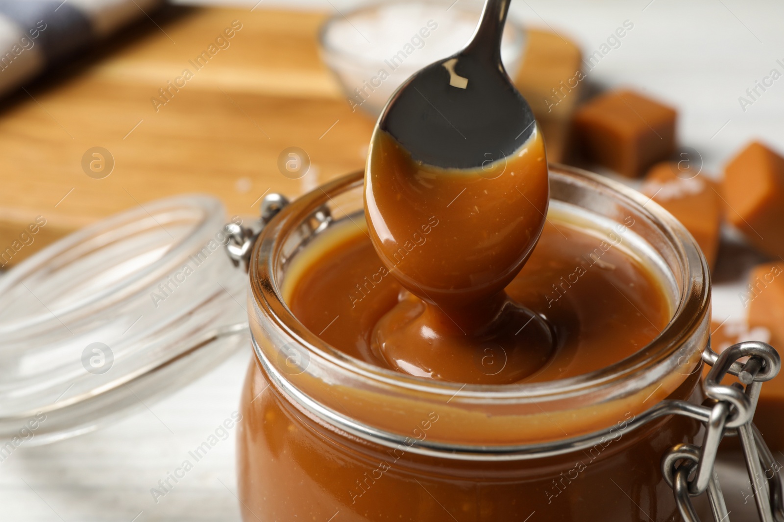 Photo of Taking yummy salted caramel with spoon from glass jar at table, closeup