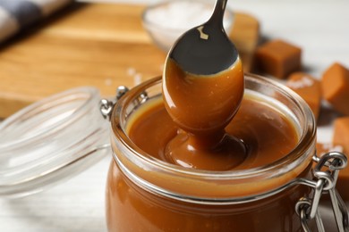 Photo of Taking yummy salted caramel with spoon from glass jar at table, closeup