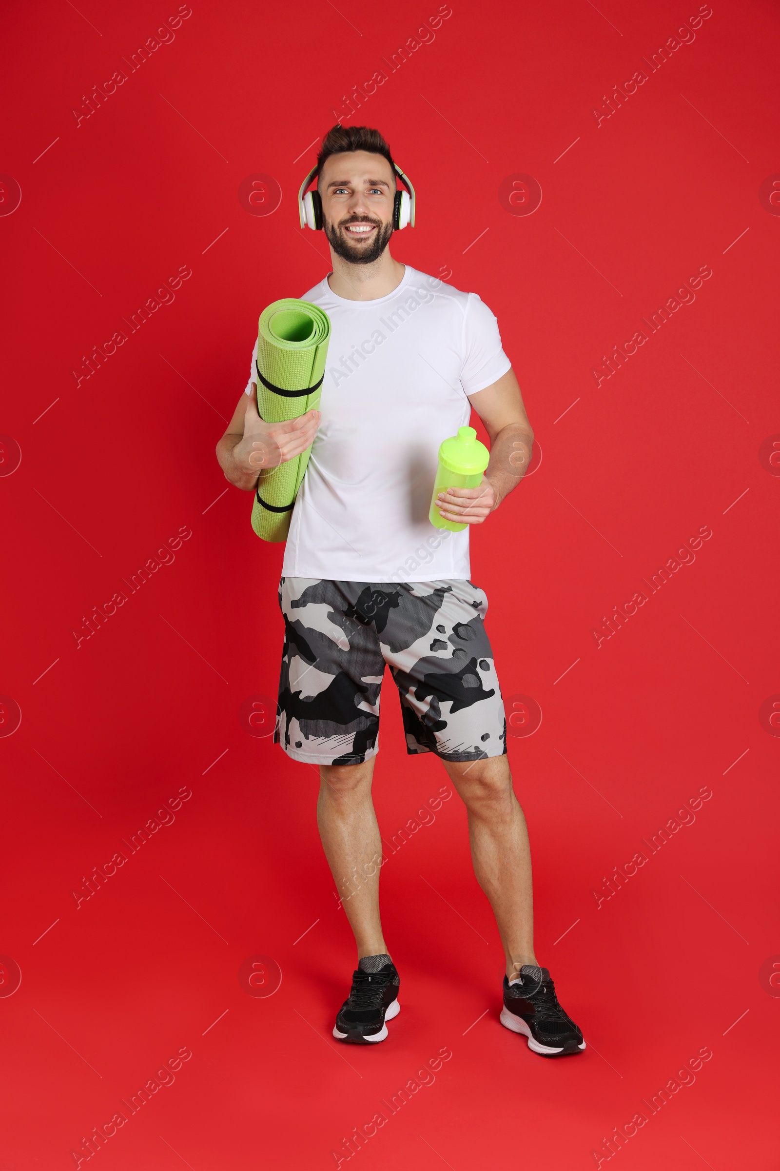 Photo of Handsome man with headphones,yoga mat and shaker on red background
