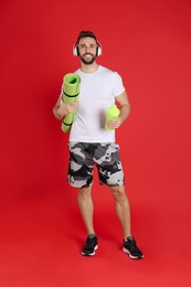 Handsome man with headphones,yoga mat and shaker on red background