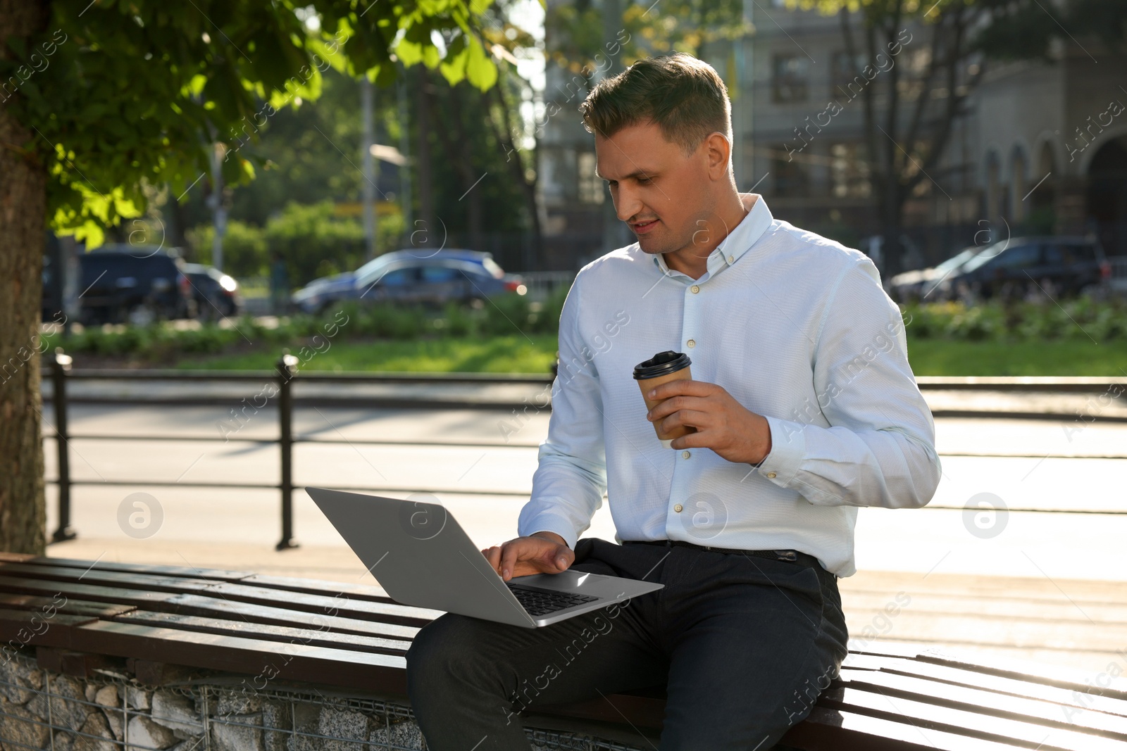 Photo of Handsome man with cup of coffee using laptop on bench outdoors