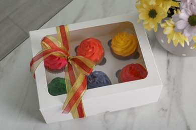 Photo of Box with delicious colorful cupcakes and beautiful flowers on white marble table